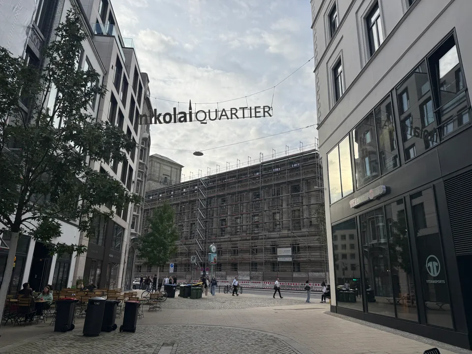 Schriftzug Nikolai Quartier über einer gepflasterten Gasse mit Café-Tischen, dahinter ein eingerüstetes historisches Gebäude