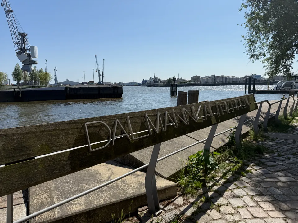 Holzbank mit Metallschriftzug Dalmannkai Treppen vor Hafenbecken, historischem Kran und Industrieanlagen am gegenüberliegenden Ufer