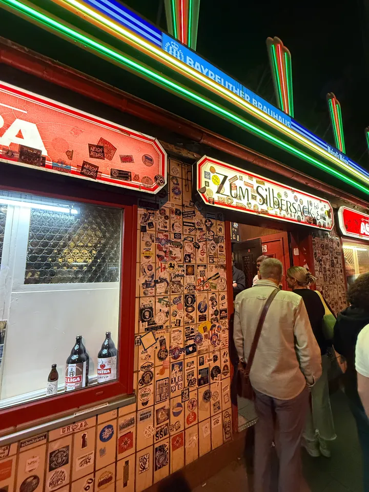 Kneipeneingang mit Schild „Zum Silbersack“, Aufkleberwand und Schaufenster mit Bierflaschen, davor anstehende Menschen