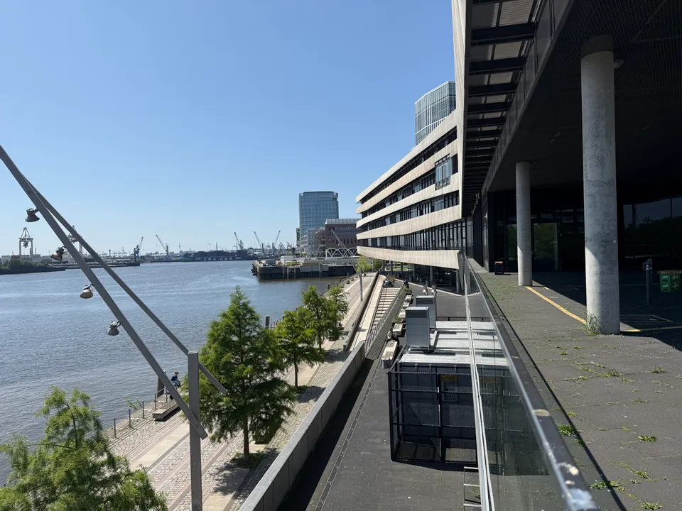 Terrasse mit Glasbrüstung neben gebänderter Gebäudefassade, links Uferpromenade, Wasser und Hafenkräne im Hintergrund