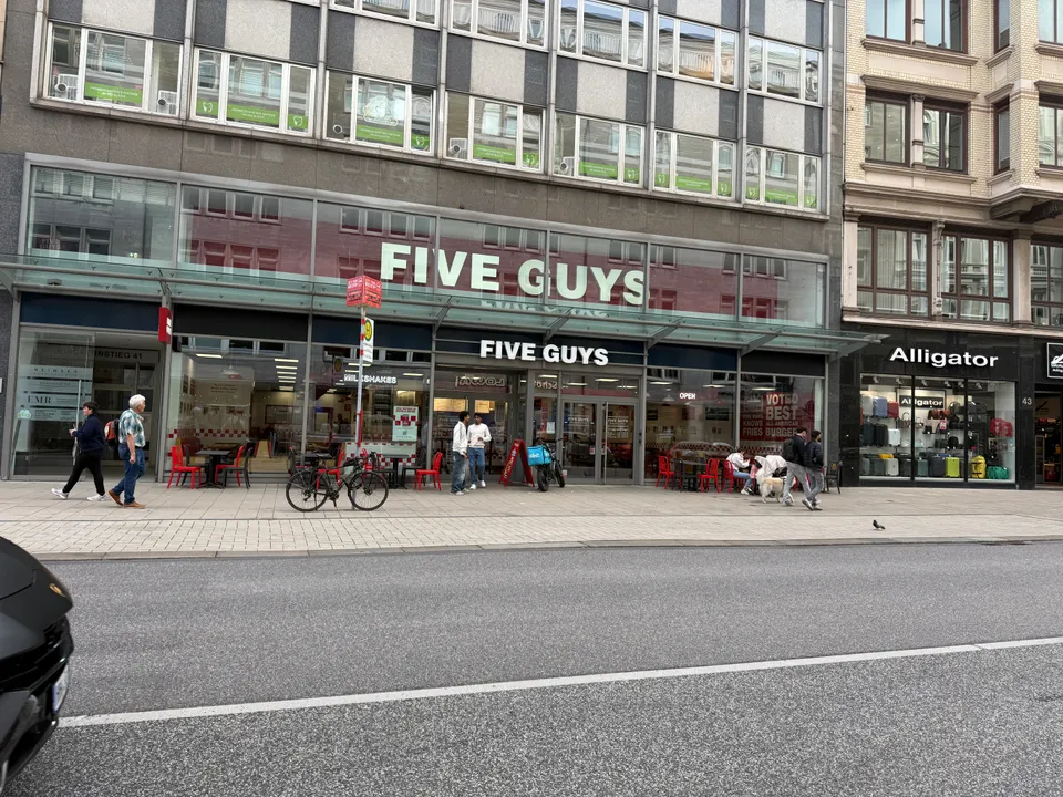 Fassade des Restaurants Five Guys am Jungfernstieg mit roten Außenstühlen, Passanten und Koffergeschäft Alligator daneben