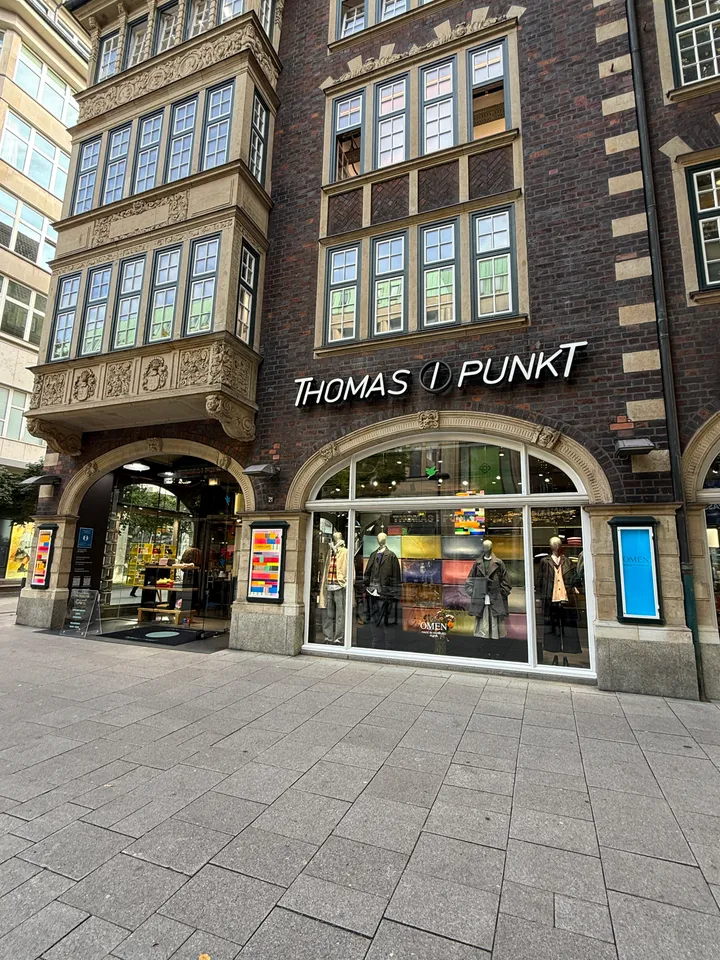 Rundbogenschaufenster mit Schaufensterpuppen unter dem Schriftzug „Thomas I Punkt“, daneben ein verzierter Sandsteinerker über dem Eingang