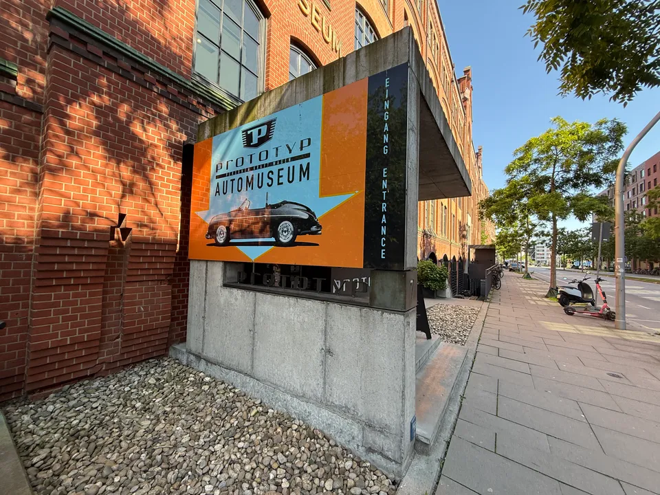 Orangefarbenes Schild Prototyp Automuseum mit Oldtimer-Cabriolet vor Backsteinfassade, daneben ein Gehweg mit Bäumen
