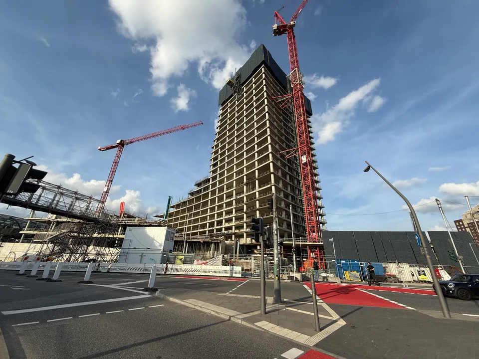 Straßenkreuzung mit roten Radwegmarkierungen vor einem Hochhausrohbau mit roten Kränen, Baucontainern und Bauzaun