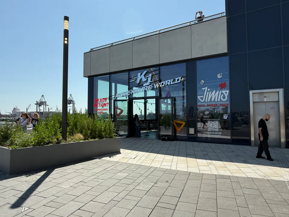 Verglaster Eingang der KJ Adventure World auf gepflastertem Platz, Hochbeet und Hafenkräne im Hintergrund