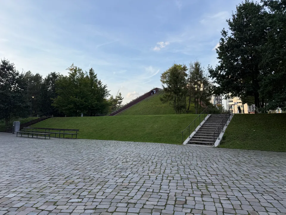 Gepflasterter Platz mit Holzbänken, Rasenterrassen, zwei Treppen und grasbewachsenem Hügel mit langer Treppe im Hintergrund