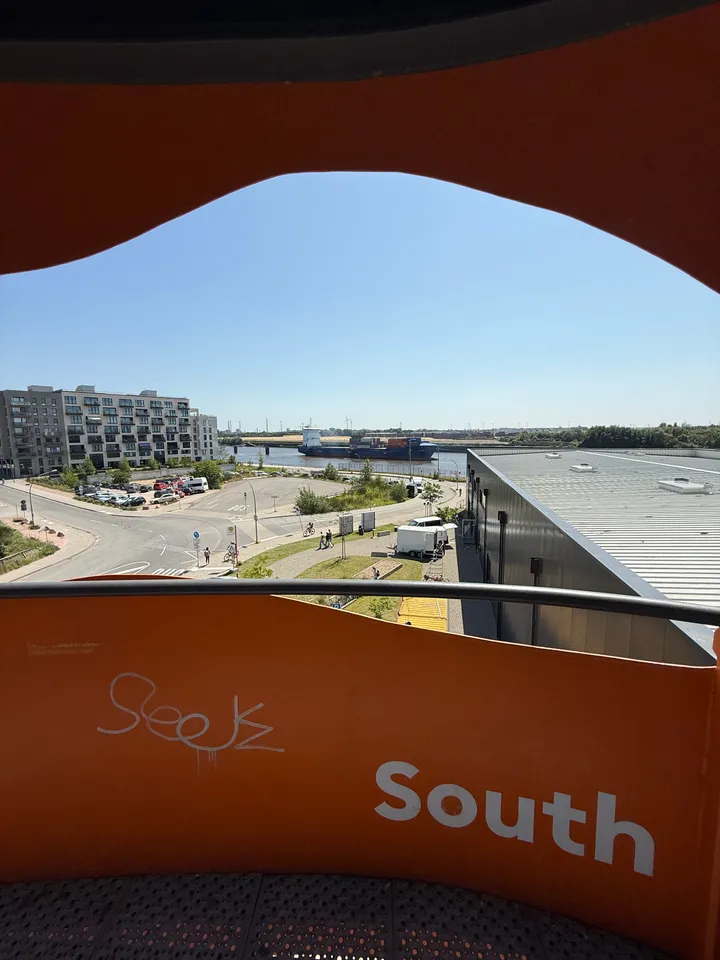 Blick nach Süden von orangefarbener Aussichtsplattform auf Straße, Wohnblock und Containerschiff auf dem Fluss