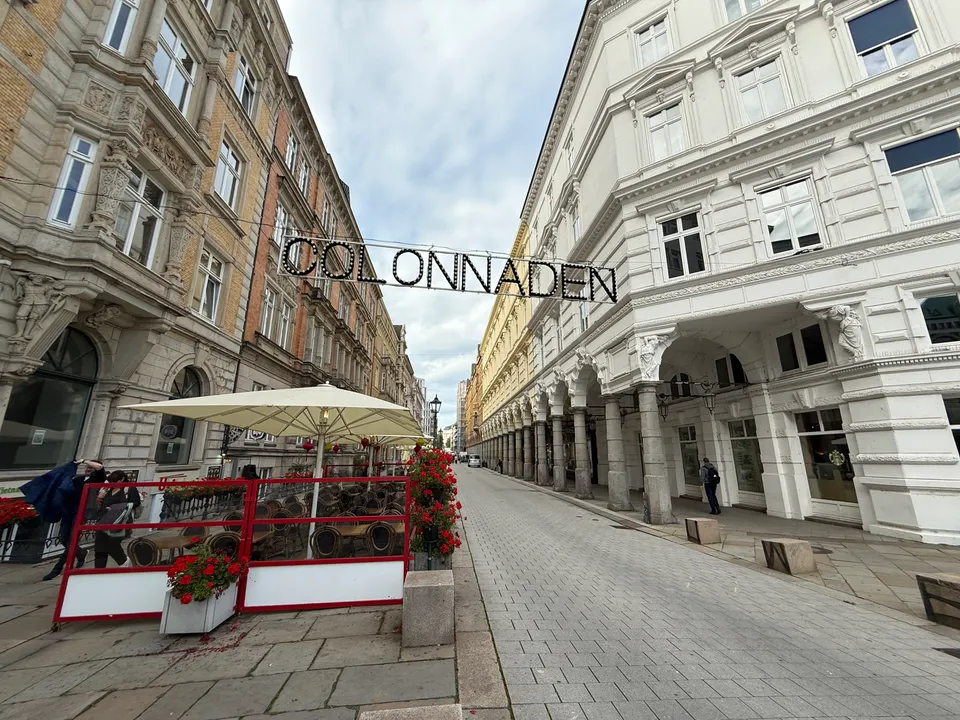 Leuchtschriftzug „COLONNADEN“ über einer gepflasterten Fußgängerstraße mit Säulenarkaden, Gründerzeitfassaden und Außengastronomie mit Sonnenschirmen