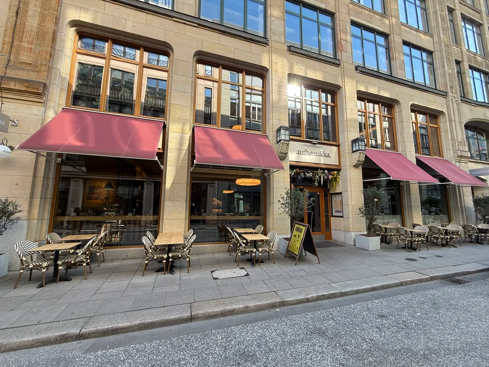 Sandsteinfassade eines Restaurants mit weinroten Markisen, Schriftzug „authentikka.“ über dem Eingang und leeren Bistrotischen auf dem Gehweg