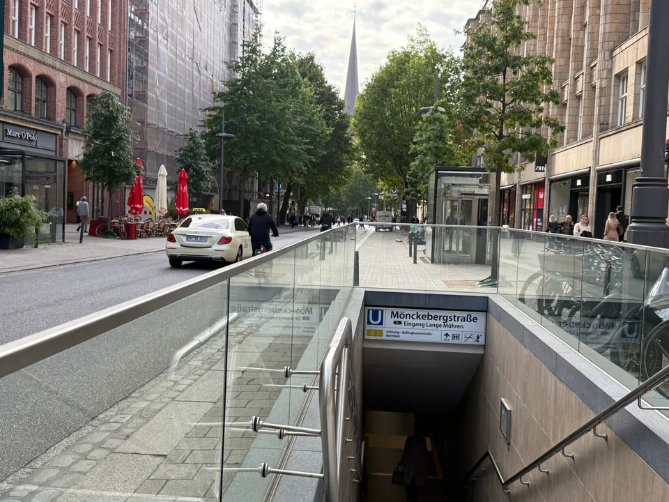 U-Bahn-Abgang Mönckebergstraße mit Glasbrüstung und Stationsschild, dahinter Straße mit Taxi, Bäumen und Kirchturm