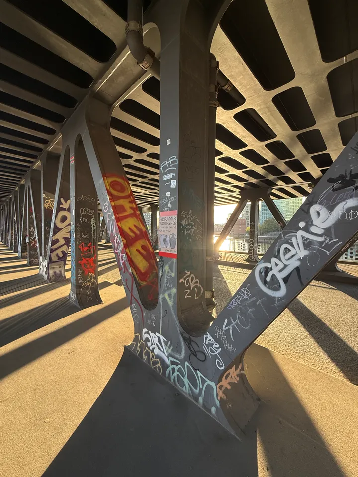 V-förmige Stahlstütze mit Graffiti-Tags unter einer Brücke, Gegenlicht und weitere Stützen im Hintergrund