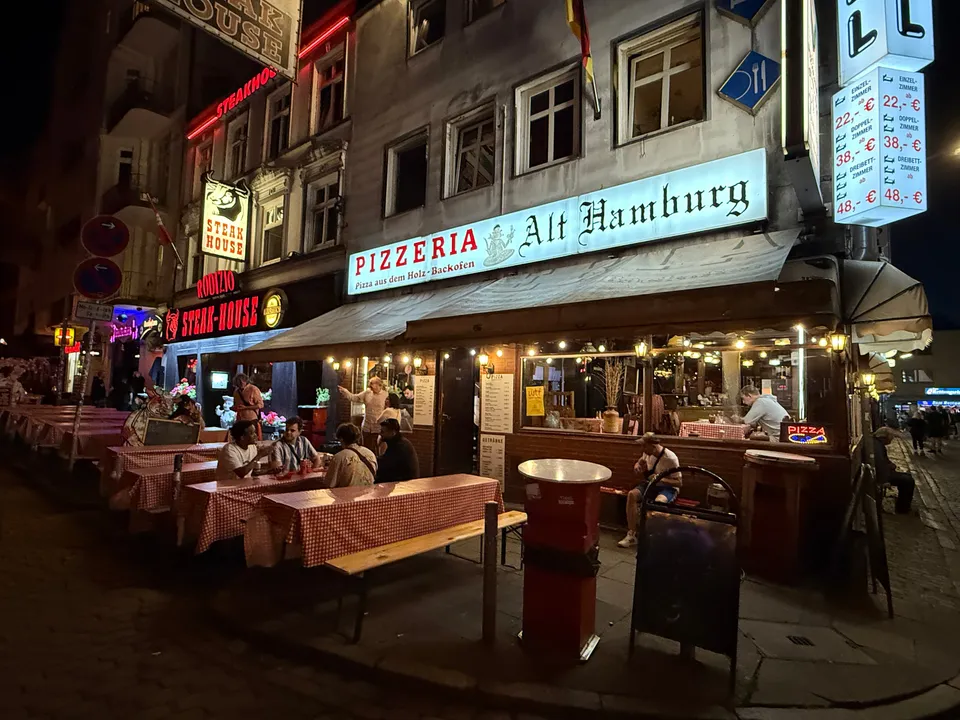 Pizzeria Alt Hamburg bei Nacht mit Außentischen, karierten Tischdecken und Steak-House-Leuchtreklame nebenan
