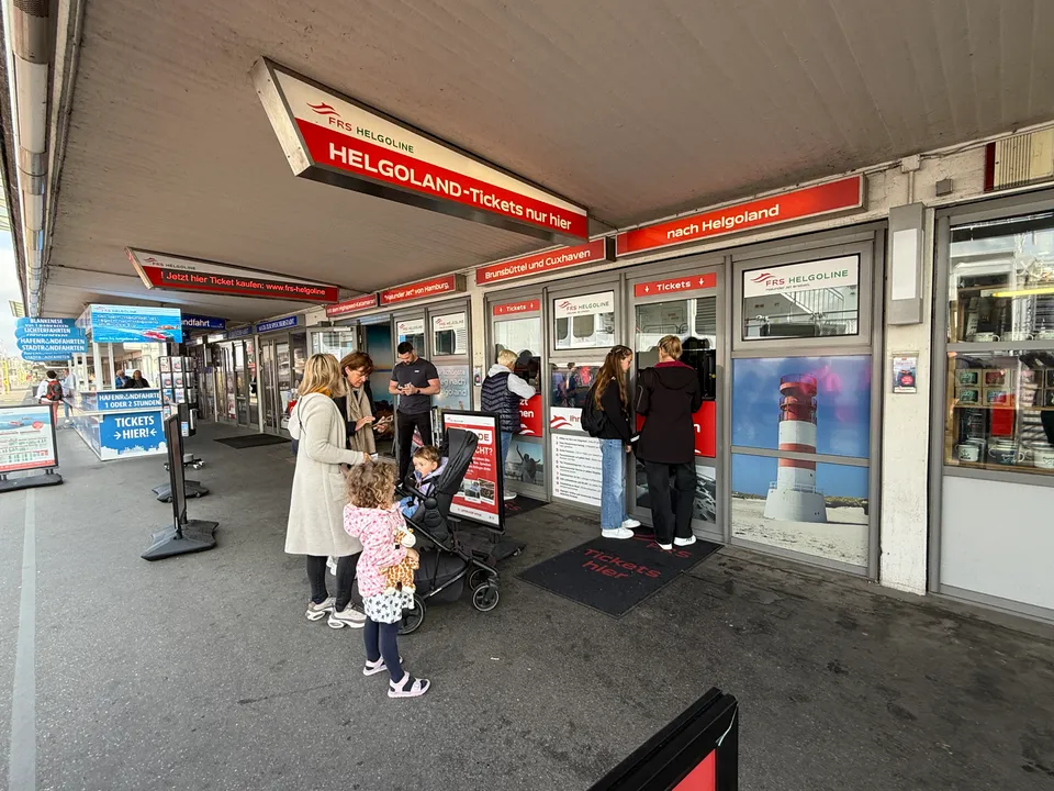 Ticketschalter der FRS Helgoline unter einem Vordach, davor wartende Menschen mit Kinderwagen und rote Helgoland-Schilder