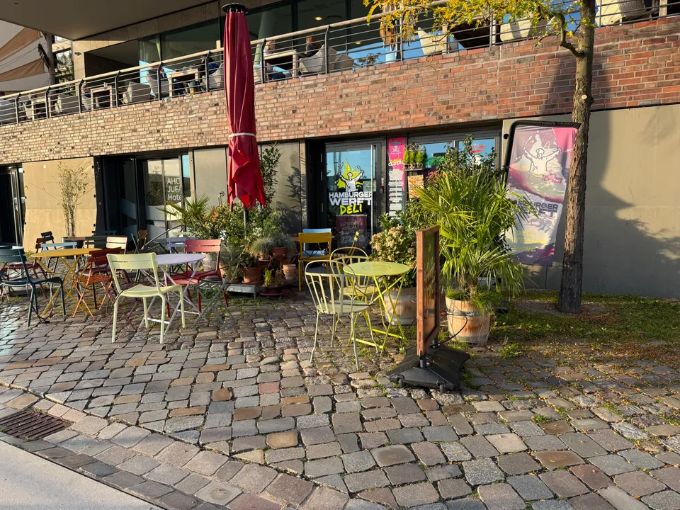 Außenbereich mit bunten Bistrostühlen, Pflanzen und rotem Sonnenschirm vor dem Hamburger Werft Deli im Klinkerbau