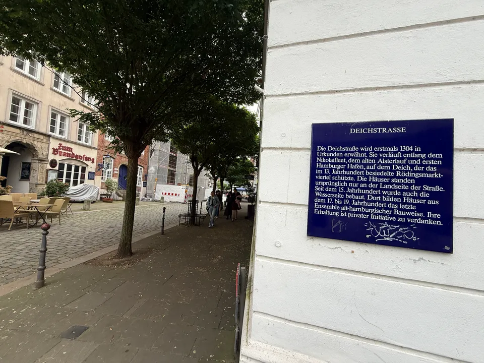 Blaue Informationstafel zur Geschichte der Deichstraße an weißer Hauswand, daneben baumbestandener Gehweg und historische Häuser