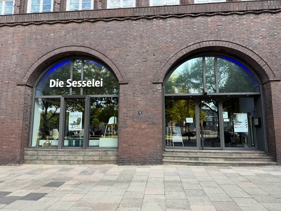 Backsteinfassade mit zwei Rundbogenfenstern, Schriftzug „Die Sesselei“ und ausgestellten Sesseln hinter Glas