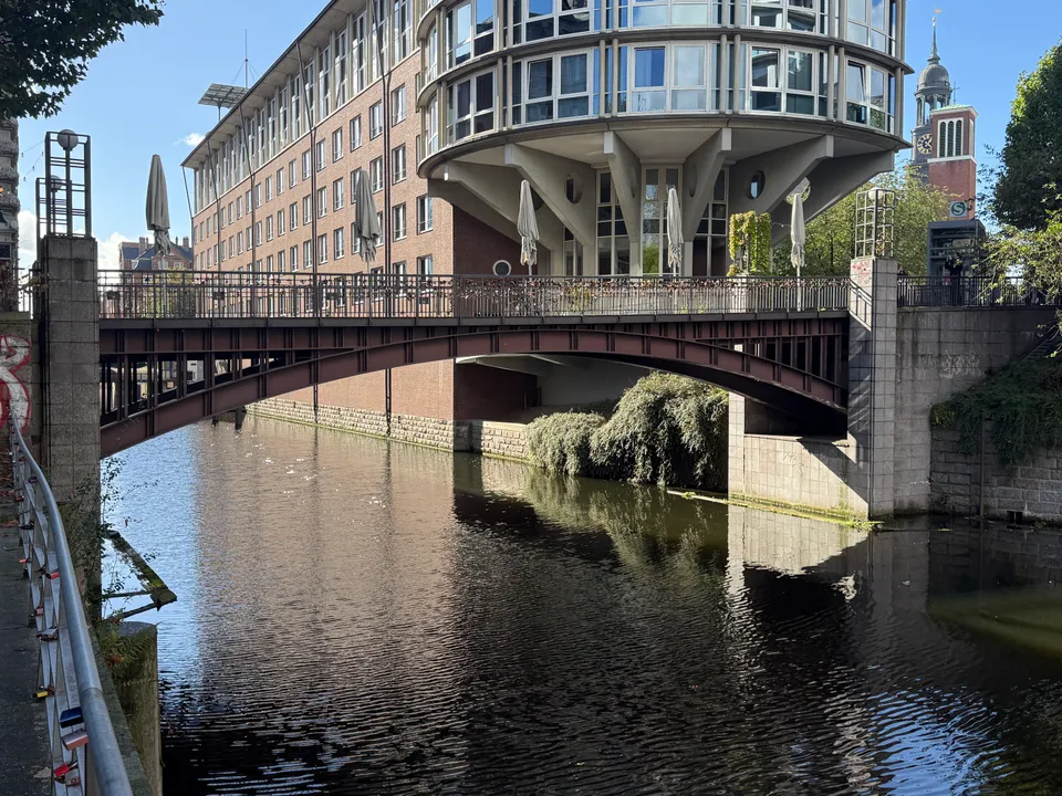 Stahlbogenbrücke über einen Fleet, dahinter ein Klinkerbau mit runder Glasfront und ein Kirchturm mit Uhr