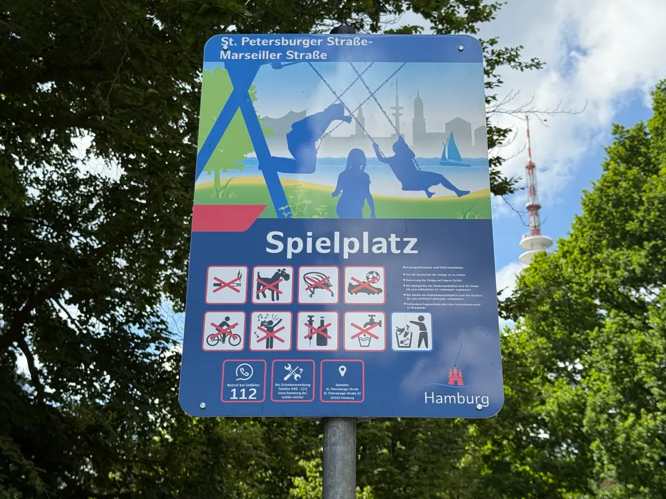 Blaues Hamburger Spielplatzschild mit Verbotssymbolen vor Bäumen, im Hintergrund der Fernsehturm
