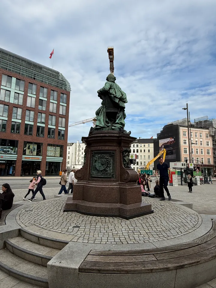 Rückansicht des Lessing-Denkmals auf rundem Podest, dahinter Backsteinbau, Baukran und Altbauten am Gänsemarkt