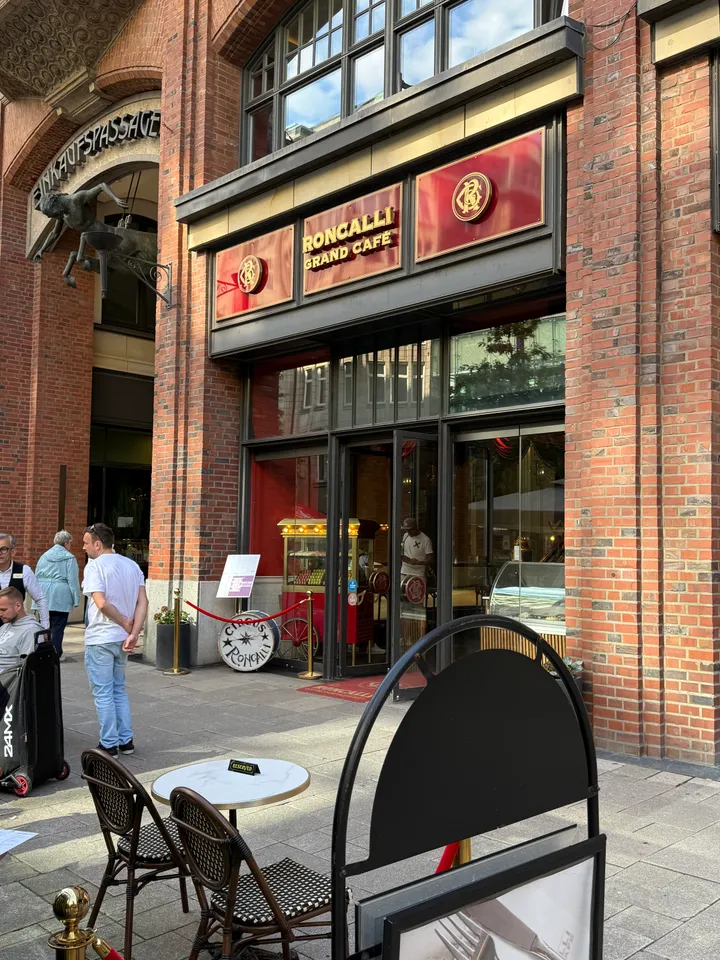 Eingang des Roncalli Grand Café mit roten Schildern, Backsteinfassade, Bistrotisch und Stühlen im Vordergrund