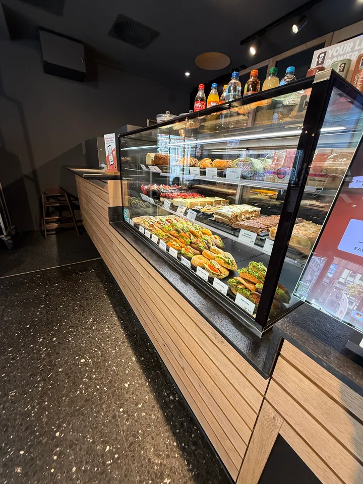 Beleuchtete Theken-Vitrine mit belegten Brötchen, Croissants und Kuchen, darauf Getränkeflaschen, vor Holzverkleidung