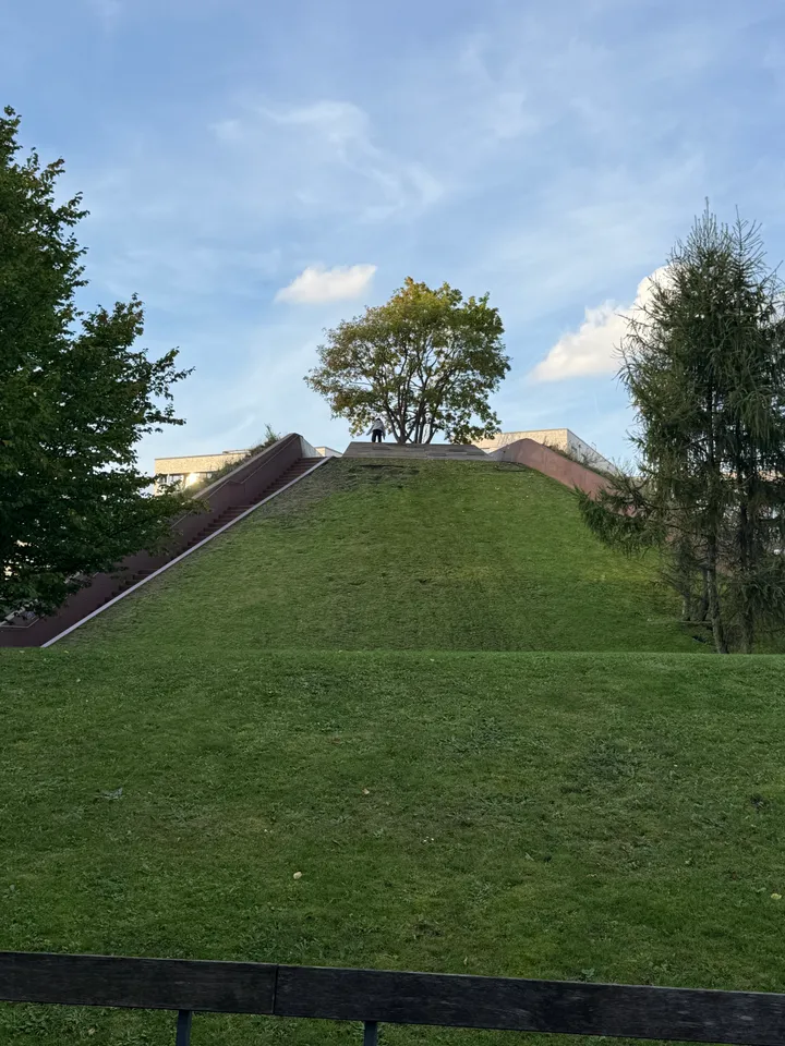 Pyramidenförmiger Grashügel mit seitlicher Treppe und einzelnem Baum auf dem Gipfel unter blauem Himmel