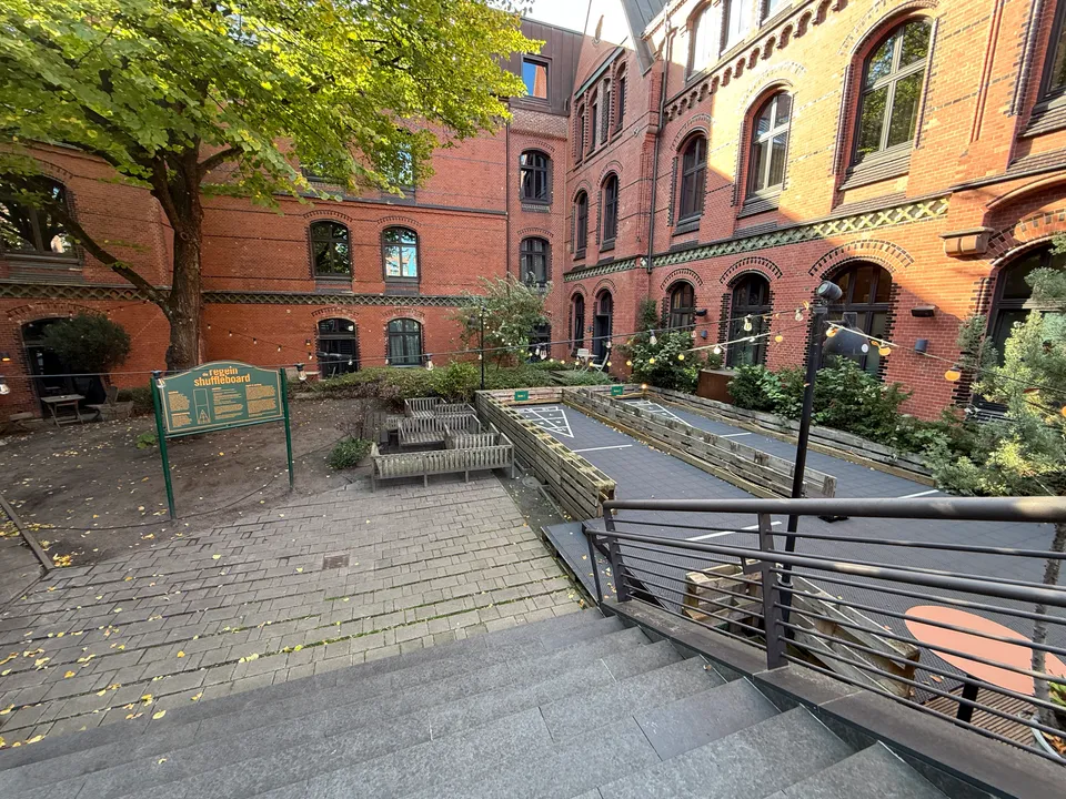 Treppe hinab in einen Backstein-Innenhof mit Shuffleboard-Bahnen, Regeltafel, Holzbänken und großem Laubbaum