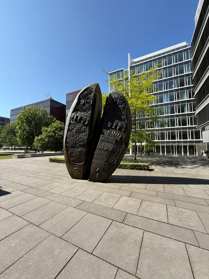 Gepflasterter Platz mit bronzener Kaffeebohnen-Skulptur zwischen modernen Glas-Bürogebäuden unter blauem Himmel