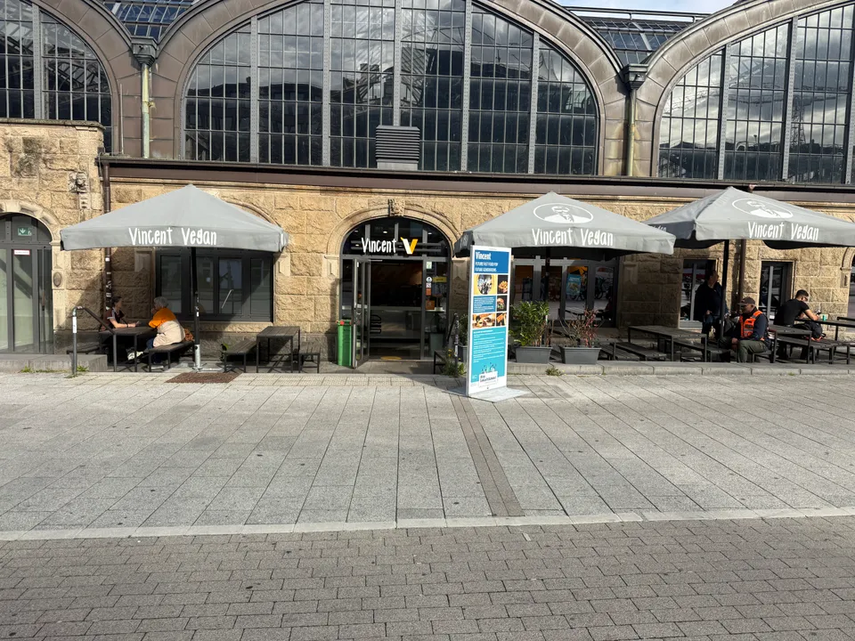 Veganes Schnellrestaurant mit grauen Sonnenschirmen und Außensitzplätzen vor der Sandsteinfassade eines Bahnhofsgebäudes mit Bogenfenstern
