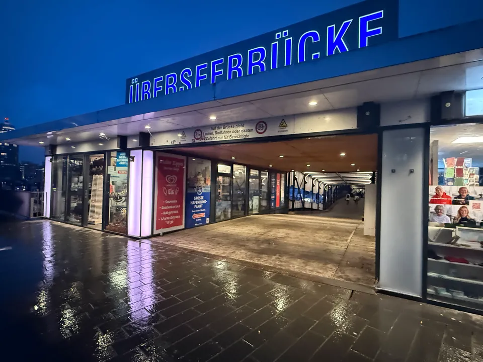 Beleuchteter Eingang der Überseebrücke mit blauem Schriftzug, Ladenfronten und nassem Pflaster in der Abenddämmerung