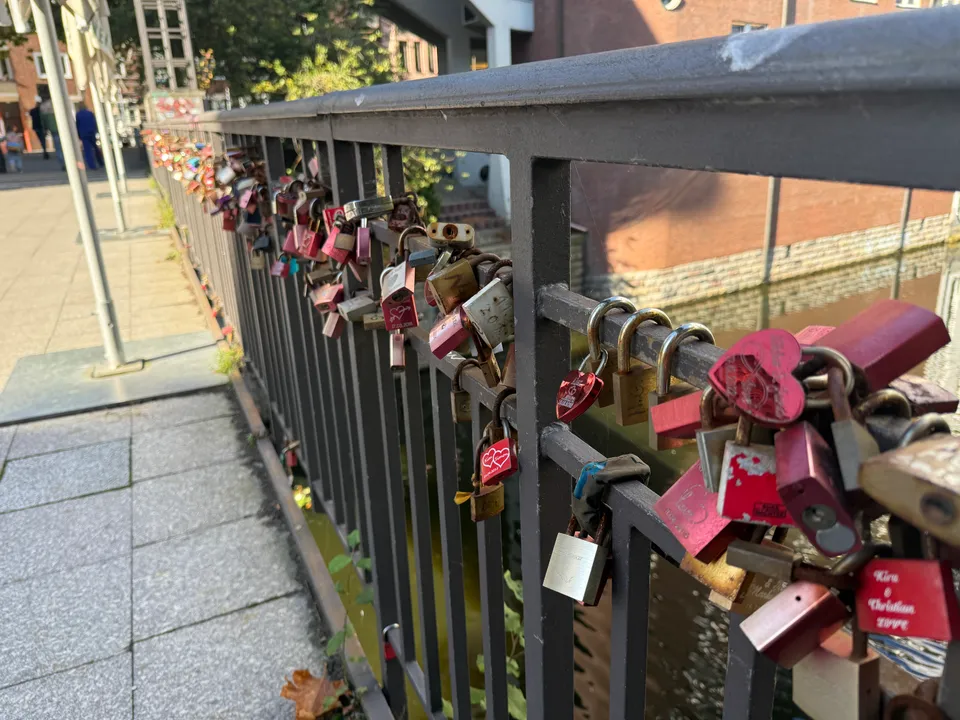 Brückengeländer mit vielen roten und messingfarbenen Liebesschlössern, daneben ein Gehweg und Wasser darunter