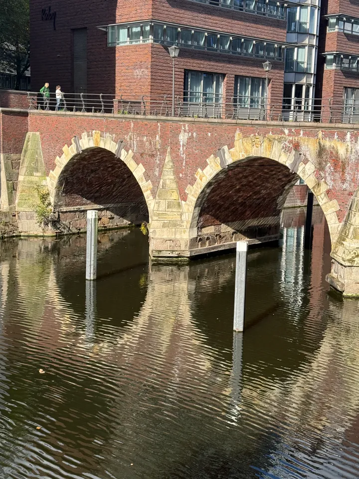 Zwei Sandsteinbögen einer Backsteinbrücke über dunklem Fleetwasser, davor zwei weiße Pfähle, dahinter ein Bürogebäude