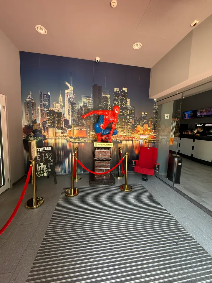 Foyer mit Spider-Man-Figur auf Backsteinsockel vor Skyline-Tapete, Absperrkordeln und rotem Kinosessel