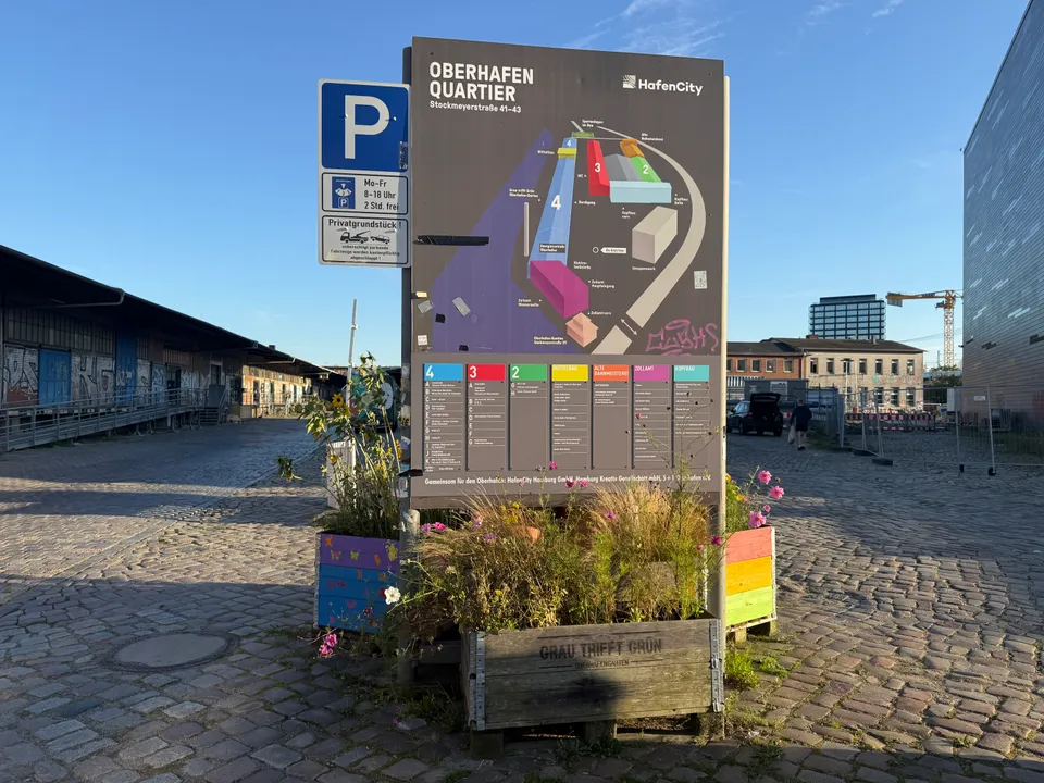Orientierungstafel des Oberhafenquartiers mit Lageplan und Parkschild, davor bepflanzte Holzkisten auf einem Platz mit Kopfsteinpflaster