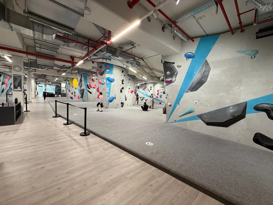 Große Boulderhalle mit grauen Kletterwänden, bunten Griffen, grauen Weichbodenmatten und freiliegenden roten Rohren an der Decke