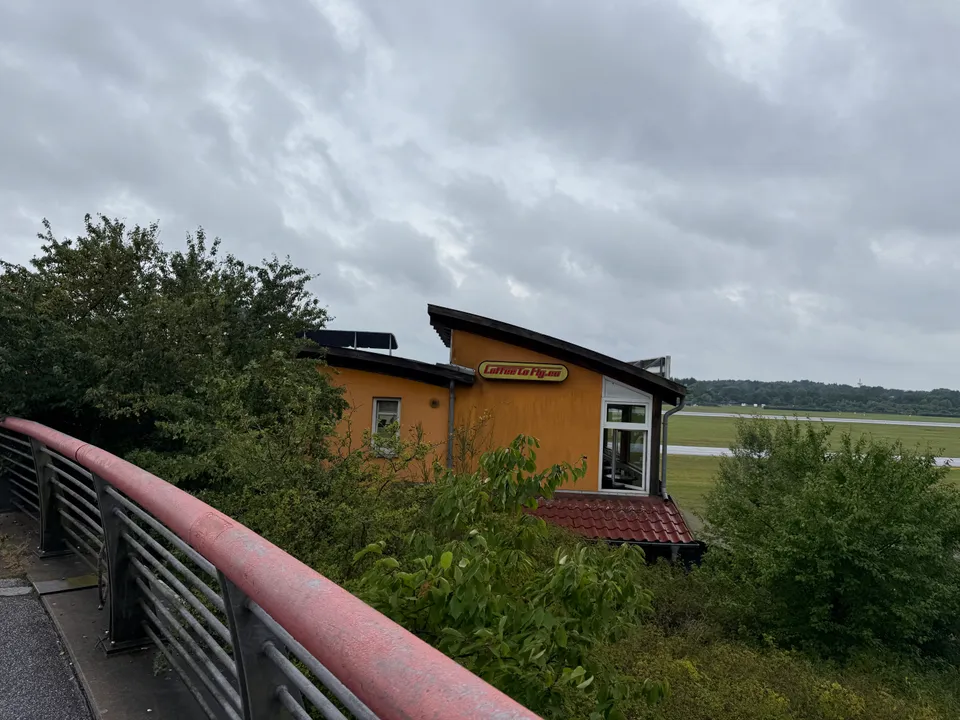Orangefarbenes Café-Gebäude mit Schild „Coffee to Fly“ hinter Büschen, daneben Brückengeländer und Flughafengelände