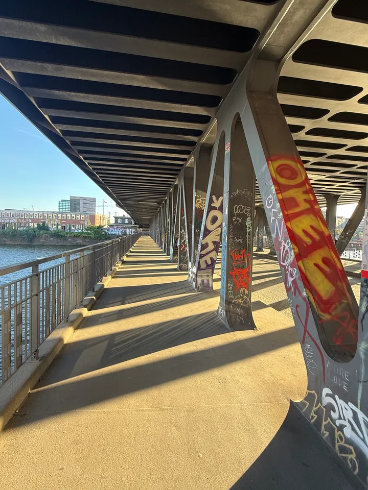 Fußweg unter einer Stahlbrücke, links Geländer und Wasser, rechts V-Stützen mit bunten Graffiti