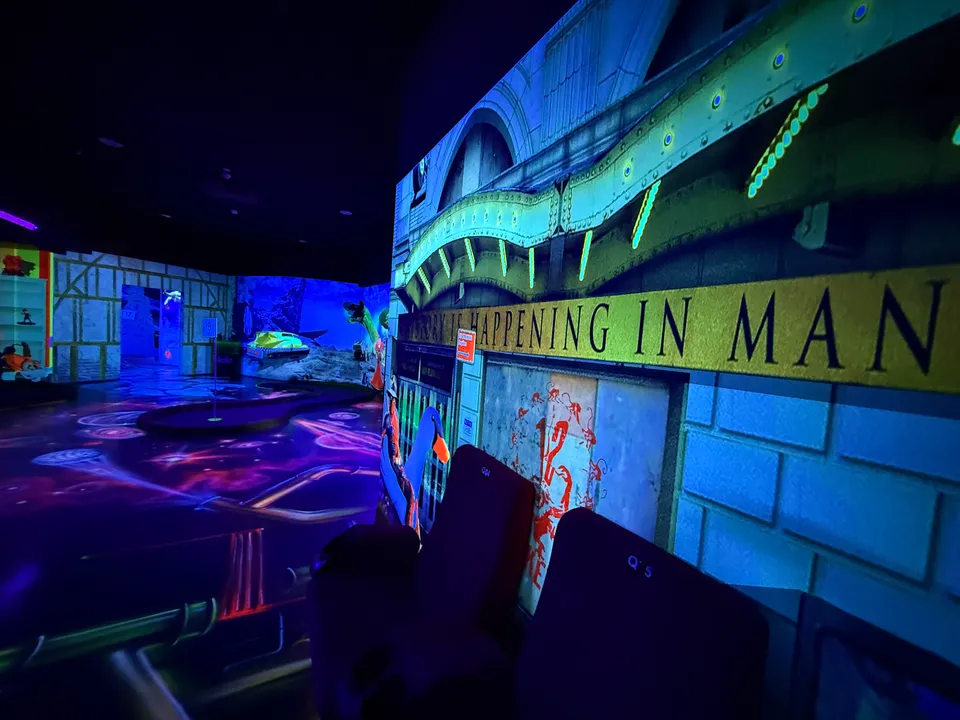 Schwarzlicht-Minigolfhalle mit Filmkulisse, Kinositzen und leuchtendem Boden, im Hintergrund eine Bahn mit Fahne