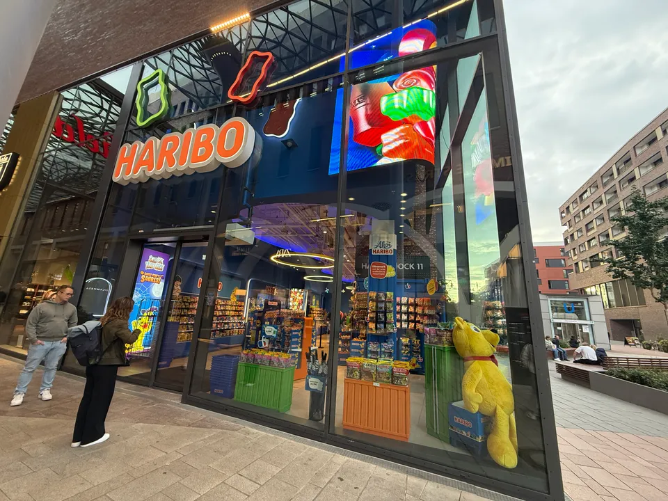 Glasfront des HARIBO-Ladens mit orangefarbenem Schriftzug, leuchtenden Goldbären-Figuren, Warenregalen und großem gelbem Plüschbären im Schaufenster