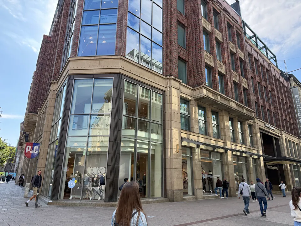 Eckgebäude mit Klinkerfassade und gläserner Schaufensterecke von Peek & Cloppenburg in einer Fußgängerzone mit Passanten