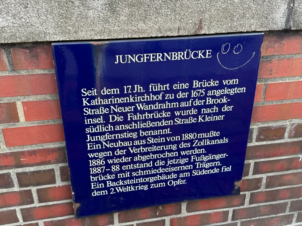 Blaue Emaille-Infotafel zur Geschichte der Jungfernbrücke an einer roten Backsteinmauer, oben rechts ein eingeritztes Smiley