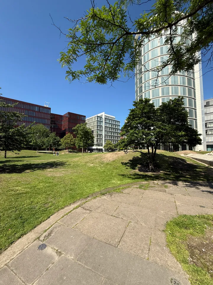 Hügelige Rasenfläche mit Bäumen und Plattenweg, im Hintergrund runder Turm und rotbraune Bürogebäude
