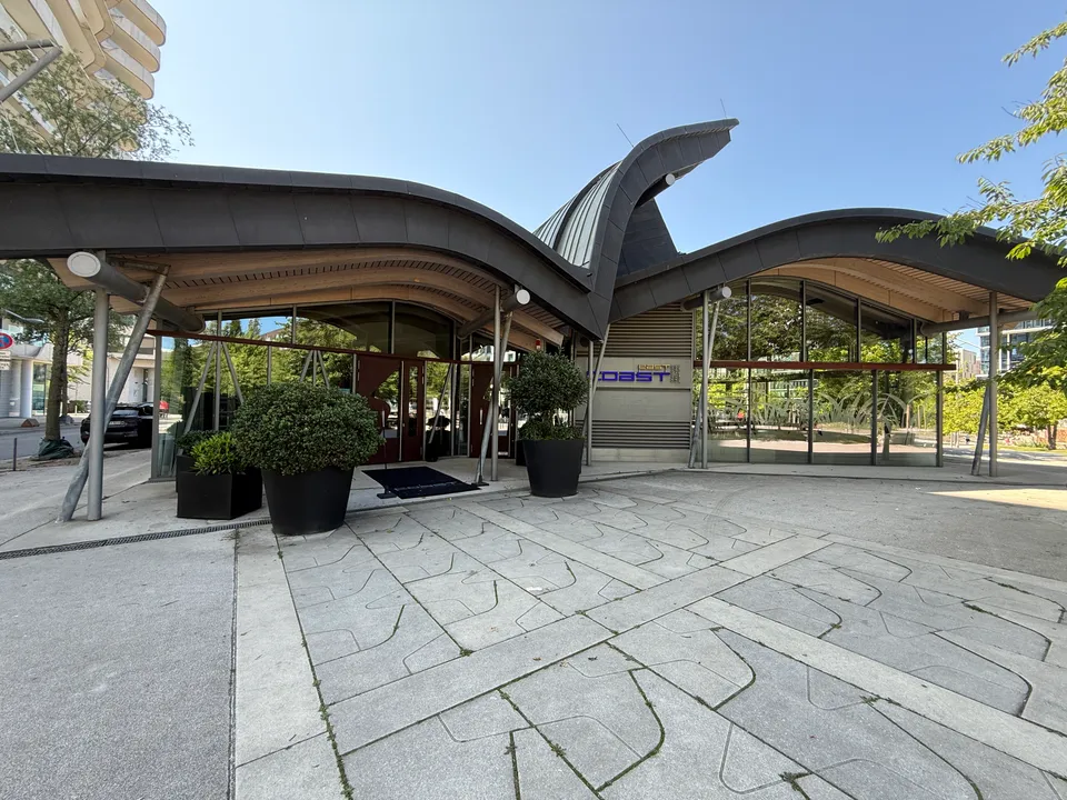 Restaurantgebäude mit wellenförmigem dunklem Dach, mittiger Dachspitze, Glasfassade und Pflanzkübeln vor dem Eingang