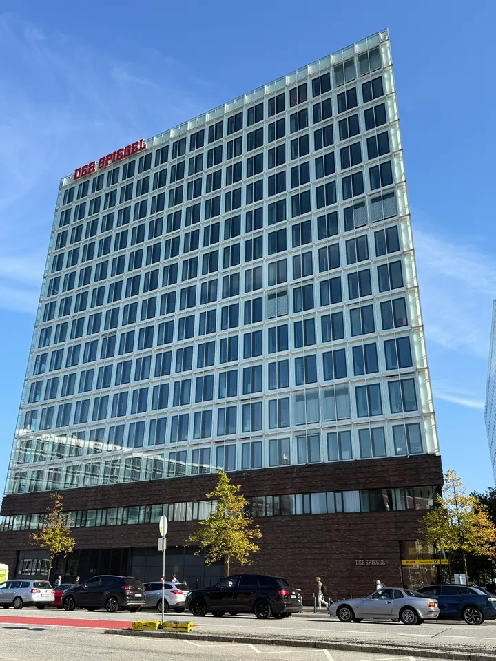 Schmale Hochhausseite des SPIEGEL-Gebäudes mit roter Dachschrift, davor Straße mit parkenden Autos und Bäumen