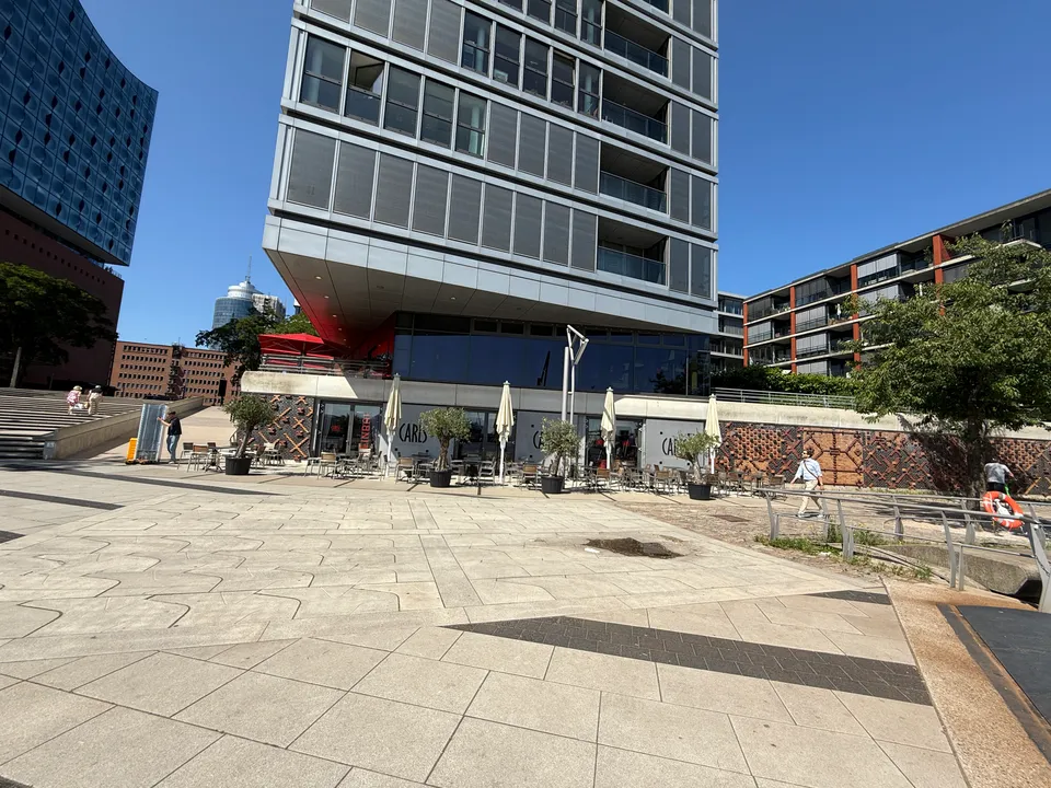 Hochhaus mit Restaurant CARLS im Erdgeschoss, Außenterrasse mit Sonnenschirmen, links angeschnitten die Elbphilharmonie, sonniger Kaiplatz