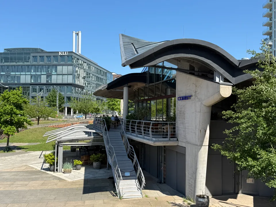 Restaurant mit spitzem geschwungenem Dach, Außentreppe und Terrasse, im Hintergrund ein gläsernes Bürogebäude mit KLU-Schriftzug
