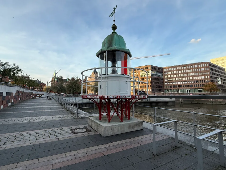 Kleine historische Leuchtfeuer-Laterne mit grüner Kupferhaube und rotem Stahlgestell an einer Hafenpromenade vor Backsteinbauten
