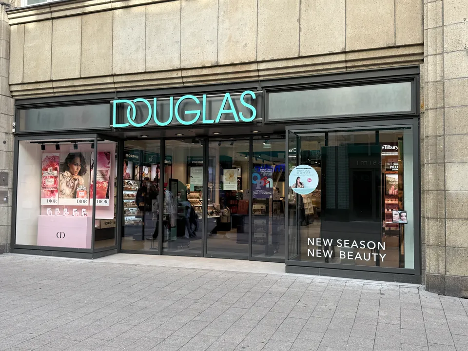 Eingang einer Douglas-Parfümerie mit türkisfarbenem Schriftzug, Dior-Schaufenster und Glasfront an gepflastertem Gehweg