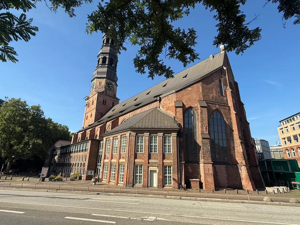 Backsteinkirche mit Turm und gotischem Chor, von Baumzweigen eingerahmt, daneben neuere Bürogebäude an einer Straße