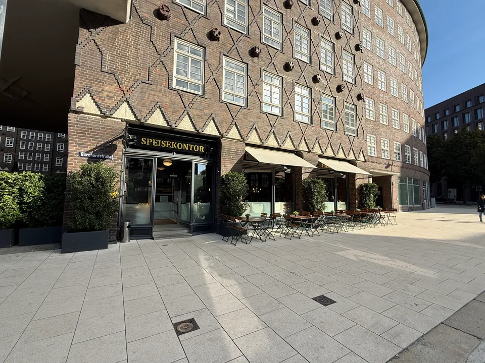 Gebogene Klinkerfassade mit Restaurant Speisekontor, Außentischen und Markisen an einem gepflasterten Platz bei Sonne